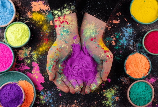 The image displays a pair of hands cupping purple powder above a black surface scattered with vibrant colored powders and several small bowls filled with different colored powders. The contrast between the vivid colors of the powder and the dark background, along with the human interaction with the materials, makes this image intriguing.